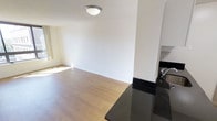 This single-floor space offers a clean, open-concept layout that seamlessly connects the kitchen and living areas. The living space features smooth light wood laminate flooring that extends throughout, complementing the bright white walls and white baseboards to create a fresh, airy feel. Natural light filters in through a window positioned along one wall, enhancing the overall brightness of the space.

The kitchen is partially visible and is distinguished by sleek dark granite countertops that provide a striking contrast against the lighter tones of the rest of the space. A stainless steel sink is also visible, suggesting functional, modern kitchen finishes. White upper cabinetry adds continuity with the overall color palette.

A flush-mount ceiling light fixture provides overhead illumination, contributing to the well-lit atmosphere of the space. The textured ceiling adds a subtle design detail throughout. The open layout between the kitchen and living area allows for flexible furniture arrangement and a sense of spaciousness. Overall, the space presents a neutral, move-in-ready aesthetic with a balanced mix of light and dark tones.