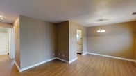 This single-floor space presents a straightforward, functional layout with a calm, neutral aesthetic throughout. The main living and dining area is finished with light-toned hardwood flooring that extends seamlessly across the space, lending warmth and visual continuity. Walls are painted in a soft taupe tone, complemented by crisp white baseboards that frame each room cleanly.

A textured popcorn ceiling adds a subtle detail overhead, while a modest chandelier with warm-toned bulbs anchors the dining area, providing a central focal point. The open layout between the living and dining zones allows for flexible furniture arrangement and creates a sense of connected, practical living.

A partial wall divides the main living area from the kitchen, which is visible through an open passageway. The kitchen features light cabinetry and countertops, maintaining a consistent, bright palette. Under-cabinet lighting provides functional illumination to the workspace.

The overall ambiance is clean, understated, and move-in ready, offering a blank canvas well-suited for personalization. The space is characterized by its simplicity, with consistent finishes and an efficient, open flow between functional areas.