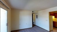 This single-floor space presents a clean, functional layout centered around a modest main room finished with wall-to-wall carpeting in a neutral tone. The walls are painted in a light, warm off-white, creating a blank canvas well-suited for personalization. The textured popcorn ceiling adds a classic finish overhead, while baseboard trim runs along the perimeter of the room, providing a simple but tidy boundary between the walls and floor.

Practical infrastructure is visibly integrated into the space, including a ceiling-mounted HVAC vent and a floor-level heating unit along one wall, ensuring year-round climate control. A recessed electrical panel is also accessible from within the room, a notable functional feature.

A doorway visible toward the back of the room leads to an adjoining area, suggesting a connected layout with additional spaces beyond the main room. The overall ambiance is straightforward and utilitarian, with minimal decorative elements, making it a practical space ready for customization. The neutral palette and consistent finishes throughout give the unit a cohesive, move-in-ready appearance.