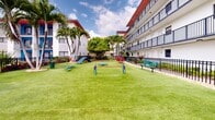 Located in Hialeah, FL, this outdoor dog park offers a well-maintained, pet-friendly recreational space designed for both dogs and their owners. The park features lush artificial turf throughout, providing a clean and consistent ground surface ideal for play and exercise year-round in Florida's climate. The space is fully enclosed by a sturdy black iron fence, ensuring a safe and secure environment for off-leash activity. A variety of colorful agility equipment is distributed across the open grounds, including a tunnel, weave poles, a ramp, and jump bars, giving dogs ample opportunity for both physical and mental stimulation. Bright blue and red benches and platforms are thoughtfully placed for owners to relax while supervising their pets. Tall palm trees add a tropical character to the space and provide natural shade. The park is nestled within the courtyard of a multi-story residential building, with balconies overlooking the grounds on multiple sides, creating a sense of enclosure and community. Decorative landscaping with colorful foliage borders portions of the fencing, adding visual appeal. The overall ambiance is energetic, colorful, and welcoming, reflecting a thoughtfully designed amenity space built to serve the needs of residents and their canine companions.