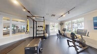 This single-story fitness facility is thoughtfully organized across several distinct areas, providing a well-rounded exercise environment. The main gym area features soaring ceilings with decorative coffered panels and pendant lighting, rubber flooring, and an abundance of natural light streaming through large arched and standard windows. Equipment includes treadmills, cable machines, strength-training stations, and cardio machines, with colorful artwork accenting the walls and a wall-mounted TV for entertainment.

A dedicated functional training room offers rubber flooring, track lighting, a power rack with TRX suspension straps, kettlebells, medicine balls, plyo boxes, and additional cardio equipment. Large windows on multiple walls flood the space with natural light, while French doors provide access to the outdoors. A full-length wall mirror enhances the sense of space.

A separate studio room, enclosed by glass partition walls and French doors, features dark flooring, mirrored walls, and a wall-mounted TV, making it suitable for group fitness or individual training sessions.

A connecting corridor with light hardwood flooring and skylight panels links the spaces and includes access to a restroom and a snack vending machine, ensuring convenience throughout the facility.