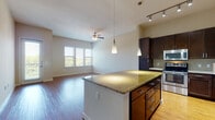 This single-floor residence is designed with an open-concept layout that seamlessly connects the kitchen and living areas, creating a cohesive and functional living space. Hardwood-style flooring runs throughout, lending warmth and consistency across each room.

The kitchen is a standout feature, showcasing rich dark cabinetry that extends to the upper walls, complemented by speckled granite countertops and a generously sized central island. A mosaic tile backsplash adds texture behind the stainless steel appliances, which include a range, built-in microwave, dishwasher, and refrigerator. Pendant lights hang above the island, while track lighting overhead provides additional illumination.

The living area is bright and open, benefiting from natural light through multiple windows that offer outdoor views. A ceiling fan adds practical comfort to the space.

A secondary room provides additional flexible living space and includes access to an exterior door. The bathroom features a tub-shower combination with white tile surround, a dark vanity with an undermount sink, and matching hardwood-style flooring, maintaining the cohesive design aesthetic found throughout the unit.