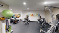 Located at 393 Porter Lake Dr in Springfield, MA, this fitness center offers a compact yet comprehensive workout facility across a single floor. The space is thoughtfully designed to maximize functionality, featuring durable rubber flooring throughout that provides both safety and comfort during exercise. Recessed lighting illuminates the room evenly, creating a bright and energizing atmosphere.

The facility is equipped with a wide range of exercise machines, including treadmills, ellipticals, stationary bikes, rowing machines, and a multi-station strength training unit. Resistance and cable machines round out the strength-training options, while stability balls, resistance bands, and a yoga mat cater to flexibility and core training needs.

A wall-mounted flat-screen TV is available for entertainment during workouts. The space is enlivened by vibrant, colorful murals depicting athletes in motion — runners and cyclists — adding dynamic visual energy to the environment. A wall-mounted climate control unit ensures year-round comfort.

French doors connect the fitness area to an adjacent laundry facility, enhancing the overall convenience of the amenity space. The clean, neutral-toned walls and well-maintained equipment make this fitness center a practical and inviting space for residents seeking a reliable on-site workout facility.