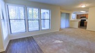 This single-floor residence offers a welcoming and well-lit interior defined by thoughtful design and practical finishes. The main living area is generously proportioned, featuring a seamless transition between hardwood-style flooring and wall-to-wall carpeting that delineates distinct functional zones within the open layout.

Natural light is a defining characteristic of this space, with a large bank of windows along one wall fitted with horizontal blinds, offering views of a private outdoor deck surrounded by mature greenery. The abundance of natural light gives the interior a warm, airy ambiance throughout the day.

The living area flows naturally toward an adjacent space, separated by vertical blinds that suggest access to additional outdoor areas or a secondary room. The soft yellow-toned walls complement the neutral flooring palette, creating a cohesive and calming aesthetic throughout.

The overall layout is open and versatile, lending itself to a variety of furniture arrangements and living styles. The interplay of light, neutral tones, and dual flooring materials gives the space a balanced character that is both functional and inviting. This residence presents a comfortable, light-filled environment well-suited for everyday living.