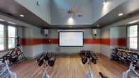 The Fit Studio is a purpose-built fitness space designed to deliver an engaging and motivating workout environment. The studio's most distinctive feature is its large projection screen mounted at the front of the room, flanked by wall-mounted speakers that create an immersive audio-visual experience ideal for guided cycling classes and virtual fitness sessions. Rows of stationary spin bikes are arranged to face the screen, allowing participants a clear sightline for instructor-led or on-screen workouts. The room's elevated ceilings add a sense of openness, while a ceiling fan ensures consistent air circulation throughout the space. The walls are accented with bold horizontal red and gray stripes, lending the studio an energetic and athletic aesthetic that complements its function. Warm wood-look flooring runs throughout, providing a clean and durable surface suited for high-traffic fitness use. Recessed lighting and overhead fixtures offer well-distributed illumination across the entire studio. The overall design balances functionality with a visually stimulating atmosphere, making the Fit Studio a well-equipped and thoughtfully designed space for group or individual cycling workouts.