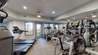 This fitness center is a comprehensively outfitted single-floor facility designed to support a wide range of workout routines. The space is finished with neutral gray walls, recessed lighting, and ceiling fans that contribute to a clean, functional ambiance. Textured carpet flooring spans throughout, providing a practical surface underfoot.

Large, multi-pane French doors and windows line the perimeter walls, flooding the interior with natural light and offering views of the surrounding outdoor area. Floor-to-ceiling mirrors are installed along multiple walls, visually expanding the space and allowing users to monitor their form during exercise.

The cardio section is well-stocked with treadmills, elliptical trainers, a stair climber, and stationary bikes from brands including Spirit and Matrix. A dedicated strength training area houses a multi-station cable machine, a full dumbbell rack ranging across multiple weight increments, and an adjustable weight bench. A separate functional training zone includes a wall-mounted rack stocked with kettlebells and medicine balls of various sizes, as well as a TRX suspension training system. A wall-mounted flat-screen television is also present, adding an entertainment element to the cardio experience. The fitness center is thoughtfully organized to allow simultaneous use of multiple equipment zones without overcrowding.
