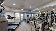 This fitness center is a comprehensively outfitted single-floor facility designed to support a wide range of workout routines. The space is finished with neutral gray walls, recessed lighting, and ceiling fans that contribute to a clean, functional ambiance. Textured carpet flooring spans throughout, providing a practical surface underfoot.

Large, multi-pane French doors and windows line the perimeter walls, flooding the interior with natural light and offering views of the surrounding outdoor area. Floor-to-ceiling mirrors are installed along multiple walls, visually expanding the space and allowing users to monitor their form during exercise.

The cardio section is well-stocked with treadmills, elliptical trainers, a stair climber, and stationary bikes from brands including Spirit and Matrix. A dedicated strength training area houses a multi-station cable machine, a full dumbbell rack ranging across multiple weight increments, and an adjustable weight bench. A separate functional training zone includes a wall-mounted rack stocked with kettlebells and medicine balls of various sizes, as well as a TRX suspension training system. A wall-mounted flat-screen television is also present, adding an entertainment element to the cardio experience. The fitness center is thoughtfully organized to allow simultaneous use of multiple equipment zones without overcrowding.