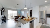 This single-floor residence showcases a thoughtfully designed open-concept layout that connects the kitchen and living areas into one cohesive, flowing space. The kitchen is anchored by a large island with a gray countertop, providing generous prep and seating space with bar-height stools. Stainless steel appliances, including a dishwasher and a sleek gooseneck faucet, complement the clean white cabinetry. Two industrial-style pendant lights hang above the island, adding a contemporary design touch.

The living area extends naturally from the kitchen, featuring light hardwood-style flooring that runs throughout. A ceiling fan enhances the open, airy feel of the space. Large sliding glass doors flood the interior with abundant natural light and lead to a balcony, framing views of the surrounding cityscape and skyline. A sectional sofa, accent chair, and curated artwork contribute to the modern, comfortable ambiance. The neutral color palette of whites and grays, accented with warm tones, gives the space a polished yet inviting character throughout.