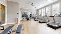 Located in Duluth, GA, this fitness center offers a comprehensive workout facility designed with both functionality and comfort in mind. The space is finished with light hardwood flooring throughout, lending a clean and inviting atmosphere that complements the neutral gray walls and crisp white crown molding and baseboards. Large windows fitted with white plantation shutters line one wall, flooding the room with abundant natural light and offering views of the surrounding greenery outside. A ceiling fan provides additional air circulation, enhancing comfort during workouts. The cardio section is well-stocked with multiple treadmills and an elliptical machine, all positioned to take advantage of the natural light. The strength training area features a multi-station cable machine, a rack of dumbbells, and a colorful assortment of kettlebells for versatile training options. A weight bench provides additional functionality for a variety of exercises. Full-length wall mirrors visually expand the space and allow users to monitor their form. Mounted flat-screen TVs keep users entertained during workouts. An adjacent room, visible through glass-paneled doors, suggests additional amenity space nearby. The overall ambiance is bright, clean, and purpose-built for an effective fitness experience.