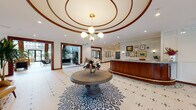 This sophisticated lobby space makes a striking first impression with its grand rotunda ceiling detail, accented by warm wood trim molding and a classic multi-globe brass chandelier at its center. The floor throughout is adorned with an intricate white mosaic penny tile pattern featuring decorative geometric inlays, adding a timeless elegance underfoot. A circular pedestal table with a fresh floral arrangement anchors the entry rotunda, welcoming visitors upon arrival through the floor-to-ceiling glass entrance doors. The curved, wood-paneled reception desk is positioned prominently, framed by framed artwork and decorative accents on the surrounding white paneled walls. Moving deeper into the space, a long corridor flanked by deep teal-painted partitioned walls with glass panels leads to a bright, open common area filled with natural light from expansive floor-to-ceiling windows. Leather seating, potted tropical plants, and pendant lighting create inviting lounge areas throughout. Rich wood-framed doorways open to additional rooms, including a stylishly furnished lounge with arched windows overlooking an outdoor area. The overall ambiance is polished and welcoming, blending classic architectural detailing with contemporary finishes.