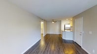 This single-floor space showcases an open-concept layout that seamlessly connects the living area to the kitchen. The floors throughout are finished in rich, warm-toned hardwood-style planks that add depth and continuity to the space. Neutral beige walls and white baseboards create a clean, versatile backdrop suited to a variety of furnishing styles.

The living area is generously proportioned, offering ample room for seating and everyday use. A ceiling fan with a light fixture is centrally positioned, providing both airflow and ambient lighting. Recessed lighting supplements the overall brightness of the space.

The kitchen is positioned to the right and features white cabinetry, a stainless steel refrigerator, and a peninsula with a dark countertop that provides additional prep and dining space. The layout encourages an easy flow between cooking and living areas.

A hallway visible in the background leads to additional rooms, hinting at further functional spaces within the unit. Overall, the space presents a well-maintained, modern interior with practical design choices and a bright, welcoming ambiance.