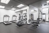 This fitness center offers a comprehensive workout environment across a single, expansive floor. The space is thoughtfully organized into distinct training zones, maximizing the available square footage for a wide range of exercise modalities. The interior features a clean, modern aesthetic with white walls, a drop tile ceiling with recessed fluorescent panel lighting, and durable speckled rubber flooring throughout — a practical surface well-suited to heavy equipment and high-intensity use.

The cardio zone is equipped with multiple treadmills, elliptical trainers, a rowing machine, and a stationary bike, providing ample options for aerobic training. Adjacent to this area, a dedicated strength training section houses a variety of selectorized machines, including cable systems and resistance equipment suited for targeted muscle training.

A functional training zone features a Rogue-branded power rack with TRX suspension straps, plyo boxes, a flat bench, and a barbell with weight plates. Nearby, a free weights area provides organized racks of dumbbells, medicine balls, and kettlebells. A punching bag is also present. Large mirrors line portions of the walls, enhancing the sense of space and supporting form-conscious training. The overall ambiance is professional and well-maintained.