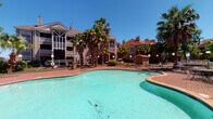 This amenity space features two distinct areas designed to support an active lifestyle. The outdoor pool area presents a generously sized, freeform swimming pool with a striking turquoise finish, encircled by a classic brick paver deck. Tall palm trees frame the surroundings, providing a resort-like atmosphere beneath open blue skies. A wrought iron fence encloses the area, with a small utility structure visible in the background alongside multi-story residential buildings.

Adjacent to the pool, the fitness center is a bright, modern space finished with neutral gray walls and textured carpet tile flooring. Floor-to-ceiling mirrors line the walls, amplifying the sense of openness and allowing users to monitor their form during workouts. Recessed lighting and a ceiling fan keep the environment well-lit and comfortable throughout. Large sliding glass doors and windows invite natural light while offering direct views of the pool area.

The gym is comprehensively equipped with multiple treadmills, stationary bikes, elliptical machines, and a full rack of dumbbells. Strength training stations, adjustable benches, cable machines, and a medicine ball rack round out the offerings. Thoughtful organizational details include wooden display shelves stocked with rolled towels and decorative plants, adding a polished, welcoming touch to the space.
