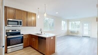 This well-designed single-floor unit offers a clean, contemporary layout centered around an open-concept kitchen and living area. The kitchen is a standout feature, showcasing rich warm-toned wood cabinetry that extends to the ceiling for ample storage, crisp white countertops, and a full suite of stainless steel appliances including a refrigerator, range, microwave, and dishwasher. A peninsula with pendant lighting overhead provides additional prep space and serves as a natural divider between the kitchen and the open living area. Light hardwood flooring runs throughout the unit, lending warmth and continuity to each space. The living area is bright and airy, with multiple windows dressed in white horizontal blinds that allow generous natural light while maintaining privacy. A glass-paneled exterior door provides direct outdoor access from the living space. The bedroom features light-toned walls, hardwood flooring, windows with greenery views, and access to a dedicated walk-in closet with built-in shelving. The bathroom is finished with a clean, all-white tub and shower combination accented by brushed bronze fixtures. The overall ambiance is fresh, neutral, and move-in ready, with a cohesive design palette that balances functionality and modern style.