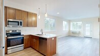 This well-designed single-floor unit offers a clean, contemporary layout centered around an open-concept kitchen and living area. The kitchen is a standout feature, showcasing rich warm-toned wood cabinetry that extends to the ceiling for ample storage, crisp white countertops, and a full suite of stainless steel appliances including a refrigerator, range, microwave, and dishwasher. A peninsula with pendant lighting overhead provides additional prep space and serves as a natural divider between the kitchen and the open living area. Light hardwood flooring runs throughout the unit, lending warmth and continuity to each space. The living area is bright and airy, with multiple windows dressed in white horizontal blinds that allow generous natural light while maintaining privacy. A glass-paneled exterior door provides direct outdoor access from the living space. The bedroom features light-toned walls, hardwood flooring, windows with greenery views, and access to a dedicated walk-in closet with built-in shelving. The bathroom is finished with a clean, all-white tub and shower combination accented by brushed bronze fixtures. The overall ambiance is fresh, neutral, and move-in ready, with a cohesive design palette that balances functionality and modern style.