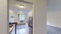 Located at Bayshore Gardens in Bay Shore, NY, this single-floor residence offers a clean, functional layout designed for comfortable everyday living. The kitchen is well-appointed with white cabinetry reaching toward the ceiling for ample storage, a stainless steel refrigerator, a gas range with stainless steel accents, a stainless steel sink, and light tile flooring. A partial wall divides the kitchen from the adjoining living area, creating a sense of separation while maintaining an open feel. The living area is generously sized, finished with plush grey carpeting and neutral white walls that lend a bright, airy atmosphere. A large window allows natural light to flow in while offering views of the surrounding greenery. Baseboard heating runs along the perimeter, ensuring year-round comfort. The bedroom mirrors the living area's clean aesthetic, featuring the same soft carpeting, neutral tones, and a window with outdoor views. The full bathroom is finished in all-white tile, including a tub-shower combination, a window for natural ventilation, a white vanity with an undermount sink, and a mirror. The overall design of the space is straightforward and well-maintained, providing a move-in ready environment in a convenient Bay Shore location.