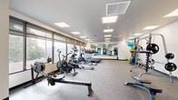 This modern fitness center is thoughtfully designed to support a comprehensive range of workout routines. The space is laid out across a single floor and is divided into distinct functional zones, providing ample room for both cardio and strength training activities.

The cardio area is lined with large windows fitted with roller shades, allowing abundant natural light to fill the room while offering views of the surrounding outdoor greenery. This section houses a row of treadmills, elliptical machines, air bikes, and rowing machines — including an Endurance rower — arranged to maximize floor space and ease of movement.

Adjacent to the cardio zone, the strength training area features a cable machine, a barbell squat rack with weight plates, a dumbbell rack with a full set of free weights, and a selection of medicine balls and exercise balls displayed on a dedicated rack. A full-wall mirror spans this section, enhancing the sense of openness and allowing users to monitor their form during exercise.

The facility features a neutral color palette of white walls, light grey ceilings, and speckled carpet flooring throughout, contributing to a clean and professional atmosphere. Recessed panel lighting in the ceiling provides consistent, even illumination across the entire space. The overall design is functional, uncluttered, and conducive to focused training.