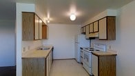 This single-floor space centers around a functional kitchen that serves as the heart of the layout. The kitchen is designed with practicality in mind, featuring an abundance of upper and lower wood-trimmed cabinetry finished in a light tone, providing generous storage throughout. A laminate countertop runs along one wall, accompanied by a stainless steel sink positioned beneath overhead lighting. On the opposite side, a white electric range with coil burners and a built-in oven sits beneath a ventilation hood, while a full-size white refrigerator stands nearby, completing the suite of essential appliances. The kitchen benefits from light-toned vinyl flooring that is easy to maintain and contributes to a clean, open feel. Overhead globe-style light fixtures provide consistent ambient illumination throughout the space. The walls are finished in a neutral white, keeping the interior bright and versatile. The overall layout is efficient and straightforward, making good use of the available space. This is a no-frills, move-in-ready environment suited for those seeking a well-equipped, functional living space with all the essential amenities in place.
