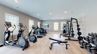 This dedicated fitness center is designed to support a full range of workout routines in a clean, modern environment. The space is finished with light hardwood-style flooring and neutral gray walls, creating a bright and motivating atmosphere. Recessed ceiling lights provide consistent illumination throughout, complemented by multiple windows fitted with blinds that allow natural light to filter in.

The cardio zone is well-stocked, featuring multiple treadmills, elliptical trainers, a stair climber, and a stationary cycle, giving users a variety of aerobic options. The strength training area is equally comprehensive, anchored by a heavy-duty Smith machine and power rack, a cable machine, and an adjustable weight bench. A full dumbbell rack holds an extensive range of free weights, and additional accessories such as resistance equipment and storage shelving are also present.

The layout is thoughtfully organized, with cardio equipment positioned near the windows to take advantage of natural light, while the strength equipment occupies the opposite side of the room. The generous floor plan allows for comfortable movement between stations. An exit door provides convenient access, and the overall design reflects a professional-grade facility suited for a broad spectrum of fitness goals.