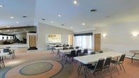 This single-floor event and gathering space is designed to accommodate a variety of functions, from community meetings to group events. The main hall is generously sized, featuring recessed lighting in a smooth white ceiling that brightens the interior evenly throughout the day. Large windows dressed with navy curtains allow natural light to fill the room, creating a welcoming and functional atmosphere. The flooring features a distinctive decorative circular pattern, adding a touch of character to the open layout. Folding tables and stackable chairs are arranged throughout the main hall, offering flexible seating configurations to suit different event needs. A pass-through kitchen window connects to a small supporting kitchen area, which includes cabinetry and countertop space — well-suited for light food preparation or catering support. The walls feature a neutral textured finish, contributing to the room's clean and practical aesthetic. Additional doors provide convenient access points, enhancing traffic flow during events. The space is well-suited for community gatherings, classes, receptions, and other organized activities, offering a straightforward and adaptable environment for a wide range of uses.