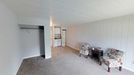 This single-floor space offers a straightforward and functional layout designed for comfortable everyday living. The main living area is finished with soft carpet flooring and features vertical panel wall detailing that lends a clean, classic character to the interior. A generously sized open closet is conveniently integrated into the living space, providing practical storage without the need for additional furniture. The neutral color palette throughout — from the light gray walls to the white paneled ceiling — creates a bright and airy atmosphere. Transitioning from the living area, the flooring shifts to light hardwood-style planks as you enter the kitchen, which is compact yet well-appointed. The kitchen includes white upper cabinetry, a countertop workspace, a stovetop, and a full-size refrigerator, offering all the essentials in an efficient footprint. The open sightline between the living area and kitchen enhances the sense of space and connectivity throughout the unit. Overall, the space presents a clean, move-in-ready environment with a simple, utilitarian design that prioritizes function and ease of use.