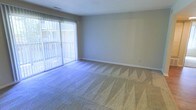This single-floor space offers a straightforward and functional layout designed for comfortable everyday living. The main living area is finished with wall-to-wall carpeting, providing a soft and welcoming underfoot feel throughout the room. Neutral gray walls create a clean, understated backdrop that complements a variety of furnishing styles and décor choices. A prominent sliding glass door fitted with vertical blinds serves as both a source of abundant natural light and a direct access point to an outdoor balcony. The balcony extends the living space outdoors, offering an area for relaxation or enjoying fresh air. The room benefits from the brightness that the large glass door provides, giving the space an airy and open ambiance even when the blinds are drawn. White baseboards accent the perimeter of the room, adding a crisp finishing detail to the neutral color palette. A ceiling vent ensures proper climate control throughout the space. Overall, the layout is open and versatile, well-suited for a range of furniture arrangements, and the connection to the outdoor balcony adds a notable sense of openness and accessibility to the exterior environment.