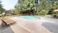 Located at 3418 SE Navigation Ln in Port Orchard, Washington, this outdoor pool facility offers a well-maintained recreational amenity set against a lush Pacific Northwest backdrop. The centerpiece is a generously sized freeform swimming pool with a kidney-shaped design, filled with clear blue-green water and equipped with dual stainless steel entry rails for easy access.

Surrounding the pool is a broad, textured aggregate concrete deck that provides ample space for lounging and circulation. A row of reclining lounge chairs lines one side of the pool, offering a dedicated area for relaxation. A patio umbrella and additional seating are positioned near the pool house, creating a shaded gathering spot.

The single-story pool house is clad in board-and-batten siding and features French doors with glass pane panels that open directly onto the pool deck. Posted signage at the entrance outlines pool rules and hours of operation, noting availability from 9:00 AM to 9:00 PM for residents and authorized guests. A first aid kit and emergency phone are mounted to the exterior wall, reflecting a commitment to safety.

The entire pool area is enclosed by a black iron fence with a gated entry, ensuring a secure environment. Towering evergreen trees frame the perimeter, providing natural privacy and a serene, forested ambiance characteristic of the greater Port Orchard region.