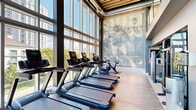 This contemporary fitness center is designed to deliver a premium workout experience across two distinct zones. The cardio area is defined by soaring floor-to-ceiling glass walls that flood the space with natural light and offer expansive views of the surrounding urban landscape. A row of commercial-grade treadmills, ellipticals, stationary bikes, and other cardio machines lines the windows, complemented by warm wood-tone flooring and a slatted wood ceiling detail that adds warmth to the industrial aesthetic. A large-scale sports mural depicting football players serves as a dramatic focal point on one wall, adding an athletic energy to the space. The strength training zone features an open, rubber-floored layout with exposed ductwork running across a dark ceiling, creating a bold industrial character. This area is outfitted with cable machines, a power rack loaded with free weights and plates, adjustable benches, a dumbbell rack, and dedicated stretching and floor exercise mats. A striking spiral staircase anchors one corner, serving as both a functional and architectural element. Together, the two zones offer a comprehensive, full-body training environment suited for a wide range of fitness goals and experience levels.
