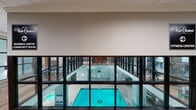 Located at 100 W Chestnut Street in Chicago's Near North Side, this amenity floor offers a comprehensive range of facilities designed to support both recreation and daily convenience. At the heart of the space is a full-sized indoor swimming pool, enclosed by floor-to-ceiling black-framed glass walls that provide visibility from the surrounding mezzanine-level lounge areas. The fitness center is expansive and well-appointed, featuring exposed brick walls, high ceilings with industrial-style ductwork, and large windows that bring in natural light from the surrounding greenery. It is equipped with a wide array of cardio machines—including treadmills, ellipticals, and stair climbers—as well as strength training equipment and free weights. Adjacent lounge and dining areas are finished with warm hardwood flooring, crown molding, and patterned upholstered seating, creating a comfortable, welcoming atmosphere. These spaces overlook the pool through glass partitions, maintaining a visual connection throughout. A dedicated game room includes a shuffleboard table and street-facing windows. The floor also includes a spacious community laundry facility with multiple washers, dryers, folding stations, and utility sinks, finished with polished concrete floors and an open ceiling.