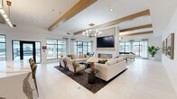 This well-appointed amenity space offers a generous, single-floor layout thoughtfully designed for social gathering and recreation. The main lounge area is anchored by an eye-catching modern chandelier and gold-accented exposed ceiling beams that lend warmth and architectural character to the otherwise crisp, white interior. Large black-framed windows line the perimeter, flooding the space with abundant natural light and offering views to the outdoors. A sleek, linear gas fireplace serves as a central focal point, framed by a textured wall panel and topped with a large flat-screen display. Comfortable seating arrangements in neutral tones are positioned throughout to encourage conversation and relaxation. Adjacent to the main lounge, a well-equipped bar area features a striking marble-topped island with seating for several guests, pendant lighting, and a full service counter with a sink, refrigeration, and a large pass-through display. The game room provides a distinct leisure zone, complete with a full-size pool table, patterned carpet flooring, artistic wall décor, and soft pendant lighting. Accessible restrooms are finished with stylish blue-green tile wainscoting and mosaic upper walls, equipped with grab bars and ADA-compliant fixtures. Throughout, the design maintains a cohesive contemporary aesthetic with curated artwork, premium finishes, and a welcoming ambiance.