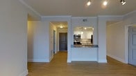 This single-floor residence offers a clean, modern aesthetic highlighted by warm hardwood flooring and crisp white walls with elegant crown molding throughout. The open-concept layout creates a seamless flow between the living area and the kitchen, which is accessible through a stylish pass-through opening framed by a granite-topped breakfast bar. The kitchen is well-equipped with stainless steel appliances, including a range, microwave, and refrigerator, complemented by white cabinetry, a decorative tile backsplash, pendant lighting, and a sink positioned at the bar counter. Track lighting above the kitchen area provides ample task illumination. A wide, well-lit hallway leads from the living space toward the entry, which features a dark, paneled front door that adds a refined contrast to the light-toned interior. Recessed vents are integrated neatly into the walls and ceiling, maintaining the clean lines of the space. The overall ambiance is bright, tidy, and move-in ready, offering a practical and attractive living environment with thoughtful design details throughout.
