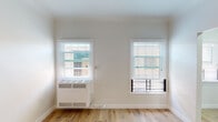 This single-floor unit showcases a clean, modern renovation with consistent design choices carried throughout each space. The living area is bright and open, featuring light-toned hardwood flooring, crisp white walls, and two windows fitted with horizontal blinds that allow for natural light and ventilation. A wall-mounted HVAC unit provides convenient climate control, while the minimalist aesthetic keeps the space feeling airy and uncluttered.

The kitchen is efficiently designed with a galley-style layout, offering ample storage through slate-gray shaker-style cabinetry on both upper and lower levels. A white quartz countertop provides a clean workspace, complemented by a deep single-basin stainless steel sink with a modern pull-down faucet. A stainless steel refrigerator and a window overlooking the exterior round out this functional and stylish kitchen.

The bathroom is a standout feature, finished with large-format marble-look porcelain tile covering the walls from floor to ceiling. A full soaking tub with chrome fixtures is set beneath a frosted privacy window, creating a refined and spa-like atmosphere. The entry area is straightforward and practical, with the same warm hardwood flooring extending throughout, anchored by a solid white door with secure hardware. Together, these spaces form a cohesive, well-appointed unit with quality finishes at every turn.