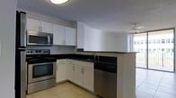 This single-floor space showcases a well-appointed kitchen designed for both functionality and style. The kitchen features crisp white cabinetry, both upper and lower, offering generous storage throughout. Black granite countertops provide a striking contrast against the white cabinets, lending a clean, contemporary aesthetic. Stainless steel appliances, including a range with oven, built-in microwave, and dishwasher, complete the modern kitchen setup. A double-basin sink is positioned along the lower cabinetry, complemented by a sleek faucet. The layout follows an L-shaped configuration, maximizing workspace efficiency. Light-toned tile flooring runs throughout, contributing to the bright and airy feel of the space. A textured ceiling adds subtle visual interest overhead. Notably, large floor-to-ceiling windows are visible adjacent to the kitchen, allowing abundant natural light to pour into the space and creating an open, connected atmosphere. The overall ambiance is clean, functional, and modern, with a neutral color palette that provides a versatile foundation for personalization.