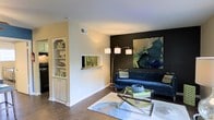 This single-story space showcases a thoughtfully designed living area that balances bold aesthetic choices with practical features. The living room serves as the focal point, highlighted by a striking dark accent wall that provides a dramatic backdrop for large-scale abstract artwork in rich blues and greens. A sleek navy velvet sofa anchors the seating area, complemented by a glass-and-metal coffee table that adds a contemporary touch. A multi-arm arc floor lamp provides ambient lighting, enhancing the room's modern character. The flooring throughout is rich hardwood, adding warmth and continuity to the space. A built-in shelving unit with cabinet storage below offers both display and organizational functionality, while a pass-through window connecting to an adjacent room adds architectural interest and openness. The walls are finished in a clean, neutral tone that contrasts effectively with the dark accent wall, contributing to a dynamic yet balanced visual composition. The overall ambiance is modern and curated, with intentional design details that create a stylish and inviting living environment.