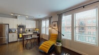 This single-floor residence offers a thoughtfully designed open-concept layout that seamlessly connects its key living spaces. An entry hallway with dark hardwood flooring and track lighting leads into the main living area, where large windows flood the space with natural light and offer views of the surrounding neighborhood. The living room features neutral tones accented by warm gold and blue décor, with a distinctive built-in media wall and a clerestory window detail that adds architectural interest above.

The kitchen is well-equipped with stainless steel appliances, including a French door refrigerator, range, microwave, and dishwasher, all complemented by granite countertops, a stainless steel backsplash, and sleek white cabinetry extending to the ceiling. A peninsula with bar seating connects the kitchen to a compact dining area.

The primary bedroom offers plush carpeting, a bold accent wall, ample dresser storage, and a clerestory window for added light. A second bedroom provides a serene retreat with soft carpet and direct access to the bathroom, which features a full soaking tub with subway tile surround and dark cabinetry.