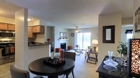 This single-floor residence presents an open-concept layout that seamlessly connects the kitchen, dining, and living areas into one cohesive living environment. The kitchen is positioned along one side, featuring wood-toned cabinetry, a dark countertop, and a breakfast bar that provides a natural transition into the dining space. A round dining table with chairs occupies the foreground, offering a casual yet functional spot for meals.

The living area is carpeted and anchored by a white-mantled fireplace, which serves as a central focal point. A ceiling fan overhead provides both airflow and ambient lighting, complementing the natural light that streams in through a large sliding glass door at the far end of the room. This door not only brightens the space but also provides direct access to an outdoor area.

Neutral wall tones and a textured ceiling contribute to a clean, understated aesthetic throughout. The open arrangement of the space promotes a sense of flow and connectivity between functional zones, making the layout feel practical and well-organized. A decorative mirror on the right wall adds depth to the overall composition.
