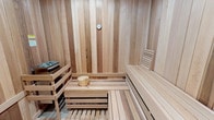 This sauna room is thoughtfully constructed with floor-to-ceiling light natural wood paneling, creating a warm and cohesive aesthetic throughout the space. The walls, ceiling, and flooring are all clad in pale, fine-grained wood — likely cedar or spruce — chosen both for its visual appeal and its practical heat-resistant properties. The consistent wood finish gives the room a clean, Scandinavian-inspired look.

The interior features tiered slatted benches along two walls, allowing users to choose their preferred heat level, as temperatures naturally increase with height. The benches are crafted from the same light wood, ensuring a uniform appearance. A traditional sauna heater, positioned in the corner, serves as the functional centerpiece of the room, accompanied by a classic wooden bucket and ladle — essential accessories for generating steam by pouring water over hot stones.

A round analog thermometer mounted on the back wall allows users to monitor interior temperature with ease. The slatted floor panels provide comfort underfoot while promoting airflow and drainage. Overall, the space offers a compact yet fully functional sauna experience with a clean, minimalist design.