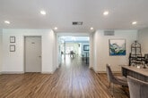 This well-appointed single-floor space offers a thoughtfully designed layout that accommodates a variety of uses, from social gatherings to everyday lounging. Warm hardwood flooring flows seamlessly throughout, unifying the distinct zones within the space.

The main lounge area is a standout, featuring dramatic vaulted ceilings with exposed white-painted beams and tongue-and-groove paneling, creating an airy and inviting atmosphere. A tiled fireplace serves as a focal point, complemented by comfortable seating and abundant natural light streaming through large windows and glass doors with views of lush greenery outside.

Adjacent to the lounge is a fully equipped kitchen, complete with dark cabinetry, granite countertops, stainless steel appliances, and a striking mosaic tile backsplash. A peninsula counter with bar-height seating provides a casual dining option.

A separate entrance corridor leads into a well-lit reception or flexible-use area with recessed lighting, clean white walls, modern artwork, and a workstation setup. This zone opens further into an additional gathering room featuring soaring ceilings with clerestory windows, high-top tables with bar stools, and a ceiling fan — ideal for casual meetings or social interaction.

The overall ambiance is clean, modern, and welcoming, with consistent finishes and a bright, open feel throughout.