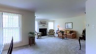 This single-floor residence offers a traditionally styled interior that emphasizes comfort and warmth. The heart of the home is a spacious living room anchored by a classic wood-mantled fireplace, which serves as a natural focal point for the space. Large windows dressed with vertical blinds allow generous amounts of natural light to filter in, brightening the neutral, light-toned walls and plush wall-to-wall carpeting throughout.

The living room is thoughtfully arranged with a coordinated seating ensemble that includes a cushioned sofa adorned with decorative pillows, a matching floral-patterned armchair, and an additional accent chair positioned near the fireplace — creating an intimate and conversational layout. A wood coffee table and side tables anchor the arrangement, while framed artwork on the walls and a table lamp add refined finishing touches to the decor.

Lush potted greenery placed near the windows and beside the seating area introduces a natural, organic element that complements the warm tones of the furnishings. The overall ambiance of the space is calm, welcoming, and classic in character — well-suited for both everyday living and relaxed entertaining. The single-floor layout ensures ease of movement and accessibility throughout the home.