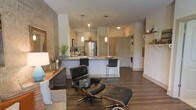 This well-appointed single-floor residence showcases an open-concept layout that seamlessly connects the living, dining, and kitchen areas, creating a spacious and cohesive atmosphere. The kitchen is a standout feature, boasting a large granite-topped island with bar seating, stainless steel appliances including a full-size refrigerator, and elegant pendant lighting that hangs above the workspace. Crisp white cabinetry and a textured accent wall add depth and character to the space. Rich, dark hardwood flooring runs throughout, grounding the interior with warmth and a polished aesthetic. The living area offers generous room for comfortable furnishings, highlighted by a classic leather lounge chair and ottoman that anchor the space with a sophisticated touch. A console table with a decorative lamp and greenery adds a refined, homey feel along one wall. Natural light filters through the space, complementing the neutral color palette of whites and warm grays. Toward the back, a dedicated in-unit laundry area provides added convenience, while interior doors lead to additional private spaces. The overall ambiance is modern yet inviting, blending clean architectural lines with carefully chosen design details.