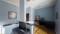 This single-floor office space offers a clean, professional environment designed for efficient daily operations. The interior is defined by a calming blue-gray wall palette complemented by crisp white crown molding and ceilings, lending the space a polished yet approachable character.

The reception or front desk area is a central feature, outfitted with sleek dark-topped counters supported by white structural columns. A desktop workstation with a monitor and keyboard is positioned at the main desk, ready for staff use. The elevated counter design creates a clear division between staff and visitor areas while maintaining an open, welcoming feel.

Along one wall, a dedicated work ledge holds a bulletin board and a printer or multifunction device, providing a practical workstation for office tasks. The warm hardwood flooring throughout adds richness and contrast to the cool wall tones, while streams of natural light cast across the floor from nearby windows, brightening the interior considerably.

Structural columns integrate seamlessly into the layout, adding an architectural element without disrupting workflow. A doorway visible in the background suggests additional rooms or spaces beyond the main reception area. Overhead, flush-mounted lighting fixtures supplement the natural light, ensuring the workspace remains well-illuminated throughout the day. Overall, this space balances functionality with a tidy, professional aesthetic.