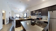 This single-floor residence showcases a thoughtfully designed open-concept layout that connects the kitchen, dining, and living areas in one cohesive, flowing space. The kitchen is a standout feature, equipped with rich dark espresso cabinetry, light stone countertops, and a full suite of stainless steel appliances including a range and over-the-range microwave. A large central island with a smooth countertop provides generous workspace and casual seating, while a secondary peninsula with an undermount sink adds further functionality. Pendant lighting hangs elegantly above both the island and peninsula, complementing the vaulted ceiling that rises gracefully over the open living area and lends an airy, expansive feel to the home. The living room benefits from natural light streaming in through multiple windows and features a ceiling fan, creating a comfortable and well-lit environment. The dining area, visible in the background, is bathed in natural light from a dedicated window, making it a welcoming spot for gatherings. Durable wood-look flooring runs throughout, tying the spaces together with a warm, contemporary aesthetic. The overall ambiance is modern, clean, and inviting.