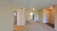 This single-floor space centers around a generously sized living area finished with wall-to-wall carpet in a warm beige tone, contributing to a clean, understated aesthetic. The walls are painted in a soft, neutral cream color throughout, creating a bright and cohesive feel across the space. A flush-mount ceiling light fixture provides central illumination, while a textured ceiling adds subtle visual depth overhead. A visible HVAC vent ensures climate control is integrated into the layout.

An open passageway connects the living area directly to the kitchen, which is partially visible from the main room. The kitchen features what appears to be light-toned cabinetry, a window that allows natural light to filter in, and a dark refrigerator, offering a functional layout. To the left of the living area, a hallway leads to additional rooms, including what appears to be a closet or utility area with a door visible at its end. A second door on the right side of the living space provides access to another room. White baseboards trim the perimeter of the space, adding a clean finishing detail throughout.