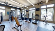 This fitness center is designed to offer a comprehensive workout experience in a bright and welcoming environment. The space is anchored by large, multi-pane windows that line the walls, flooding the interior with natural light and offering views of the surrounding outdoor landscape, including lush greenery and neighboring architecture. The light wood-style flooring adds a clean, modern aesthetic throughout.

The cardio section is well-stocked with treadmills, elliptical trainers, and rowing-style machines, all positioned to take advantage of the natural light and outdoor views. Adjustable workout benches are conveniently placed throughout the space to support a variety of strength training exercises. A multi-station cable machine provides additional options for resistance training.

Wall-mounted flat-screen televisions are positioned at strategic points around the room, allowing members to stay entertained during their workouts. Ceiling fans installed above help maintain comfortable air circulation throughout the space. Overhead fluorescent lighting ensures the room remains well-lit regardless of the time of day. The overall ambiance is open, energetic, and functional, making this fitness center a practical and inviting space for a full range of exercise routines.