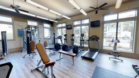 This fitness center is designed to offer a comprehensive workout experience in a bright and welcoming environment. The space is anchored by large, multi-pane windows that line the walls, flooding the interior with natural light and offering views of the surrounding outdoor landscape, including lush greenery and neighboring architecture. The light wood-style flooring adds a clean, modern aesthetic throughout.

The cardio section is well-stocked with treadmills, elliptical trainers, and rowing-style machines, all positioned to take advantage of the natural light and outdoor views. Adjustable workout benches are conveniently placed throughout the space to support a variety of strength training exercises. A multi-station cable machine provides additional options for resistance training.

Wall-mounted flat-screen televisions are positioned at strategic points around the room, allowing members to stay entertained during their workouts. Ceiling fans installed above help maintain comfortable air circulation throughout the space. Overhead fluorescent lighting ensures the room remains well-lit regardless of the time of day. The overall ambiance is open, energetic, and functional, making this fitness center a practical and inviting space for a full range of exercise routines.
