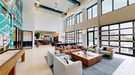 Located at 201 Pilot St in Savannah, Georgia, this expansive amenity lounge offers a thoughtfully designed, open-concept space that spans a generous single floor. The most striking architectural feature is the dramatic double-height ceiling, which floods the interior with natural light through floor-to-ceiling gridded windows that frame views of the surrounding neighborhood.

The main lounge area is anchored by a rich mix of contemporary furnishings, including a deep cognac leather sectional, soft gray accent chairs, and a substantial wood-topped coffee table, all arranged atop a large area rug. Pendant lighting with globe fixtures descends from the soaring ceiling, complementing the dark teal beam accents overhead.

A dedicated game area features a full-length wooden shuffleboard table set against a bold, large-scale abstract mural in teal, orange, and white tones, adding a strong artistic focal point to the space. Adjacent to this is a fully equipped kitchen outfitted with dark cabinetry, a large white quartz island with bar seating, stainless steel appliances, and hexagonal tile flooring. Glass doors connect the kitchen directly to an outdoor area.

A secondary lounge zone offers additional seating with woven accent chairs and a high-top bar counter, further enhancing the versatility of this well-appointed communal space.