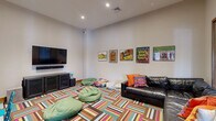 Located on Riley Road in Easton, PA, this single-room living space offers a fun and energetic environment designed for comfort and casual gathering. The room is defined by a bold, multicolored geometric patterned carpet that immediately draws the eye and sets a lively tone throughout the space.

The entertainment setup is anchored by a wall-mounted flat-screen television paired with a soundbar and a low-profile media console with cabinet storage. Seating options are varied and inviting, including a large dark leather sectional sofa adorned with vibrant throw pillows, several green bean bag chairs arranged across the floor, and a compact high-top table with teal metal chairs for more upright seating.

The walls are decorated with a curated collection of colorful framed artwork, adding a playful and creative character to the room. Natural light filters in through a window and glass-paneled double doors with wood trim, which lead to an adjacent bright space and contribute to an open, welcoming feel.

The room features neutral-toned walls that contrast beautifully with the colorful furnishings, and recessed ceiling lighting provides warm, even illumination. The overall ambiance is casual, energetic, and well-suited for relaxation, media viewing, or social interaction in a compact yet thoughtfully arranged setting.