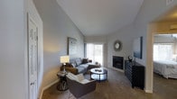 This single-floor living space showcases a thoughtfully designed interior centered around a spacious living room. The room's most striking architectural feature is its vaulted ceiling, which dramatically increases the sense of height and openness throughout the space. Neutral gray walls and carpeted flooring establish a calm, cohesive aesthetic that complements the contemporary furnishings.

A tile-surround fireplace serves as a natural focal point along one wall, providing both a functional and decorative element to the room. Adjacent to the fireplace, a dark-toned media console adds practical storage while anchoring the entertainment area. A large sliding glass door allows generous natural light to fill the space and offers a view of the outdoors, further enhancing the airy feel created by the vaulted ceiling.

The living area is furnished with a sectional sofa, accent chair, and a round coffee table, creating a comfortable and functional seating arrangement. Decorative touches, including framed wall art and a sunburst mirror, contribute to the polished, well-curated ambiance of this inviting single-level home.