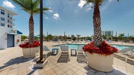 Located at 2500 SW 22nd Terrace in Fort Lauderdale, Florida, this outdoor amenity space presents a well-appointed resort-style pool deck designed for leisure and relaxation in a sunny South Florida setting. The centerpiece of the area is a generously sized swimming pool with clear, blue-tinted water, flanked on multiple sides by a broad, light-toned paver deck that provides ample room for movement and socializing. Rows of lounge chairs are arranged along the poolside, offering dedicated spaces for sunbathing and outdoor relaxation. Tall, mature palm trees are strategically positioned throughout the deck, lending a tropical character to the environment while providing natural shade. Vibrant red flowering plants in large planters add striking color accents against the crisp white façade of the multi-story residential building that frames the space. Shaded seating areas with umbrellas and cabana-style structures are available at the periphery, offering relief from the sun. Amenity structures including restroom facilities and utility access points are neatly integrated into the layout. A railed walkway connects the pool area to surrounding building access points, ensuring ease of circulation. Open views of the surrounding Fort Lauderdale skyline and lush greenery are visible beyond the perimeter, enhancing the overall outdoor experience. The deck is finished with large-format pavers that complement the clean, modern architectural aesthetic of the building.