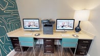 This business center is a compact yet fully functional workspace designed to meet a variety of professional needs. The room is anchored by a long, light-colored work surface accommodating two Apple iMac computers, each paired with a wireless keyboard and mouse, making simultaneous use straightforward and efficient. A Brother multifunction printer and scanner sits centrally on the desk, providing convenient print and copy access. Teal-accented bar-height chairs with sleek metal frames complement the desk's dark espresso storage drawers, lending a cohesive and modern aesthetic throughout.

One of the most striking design elements is the bold, large-scale abstract wallpaper in deep teal with gold linework, which covers select walls and adds a dramatic visual focal point to the otherwise neutral space. The remaining walls are finished in a soft, light gray, creating a balanced contrast. Wood-look flooring runs throughout, adding warmth underfoot.

Natural light enters through a window fitted with horizontal blinds, offering views of the surrounding area. A French glass door provides entry and maintains a sense of openness while visually connecting the business center to adjacent spaces. Ambient lighting from a decorative table lamp rounds out the well-considered atmosphere, making this center both practical and visually engaging.